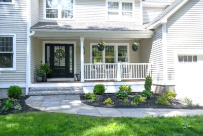 - Courtyard Concepts Front porch of a beige siding house with a black front door, white railings, and hanging planters.