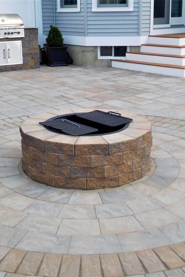 Circular stone-paver patio with a built-in stone fire pit at center and a grilling station along the back wall; multi-tiered steps lead to a doorway on the right.